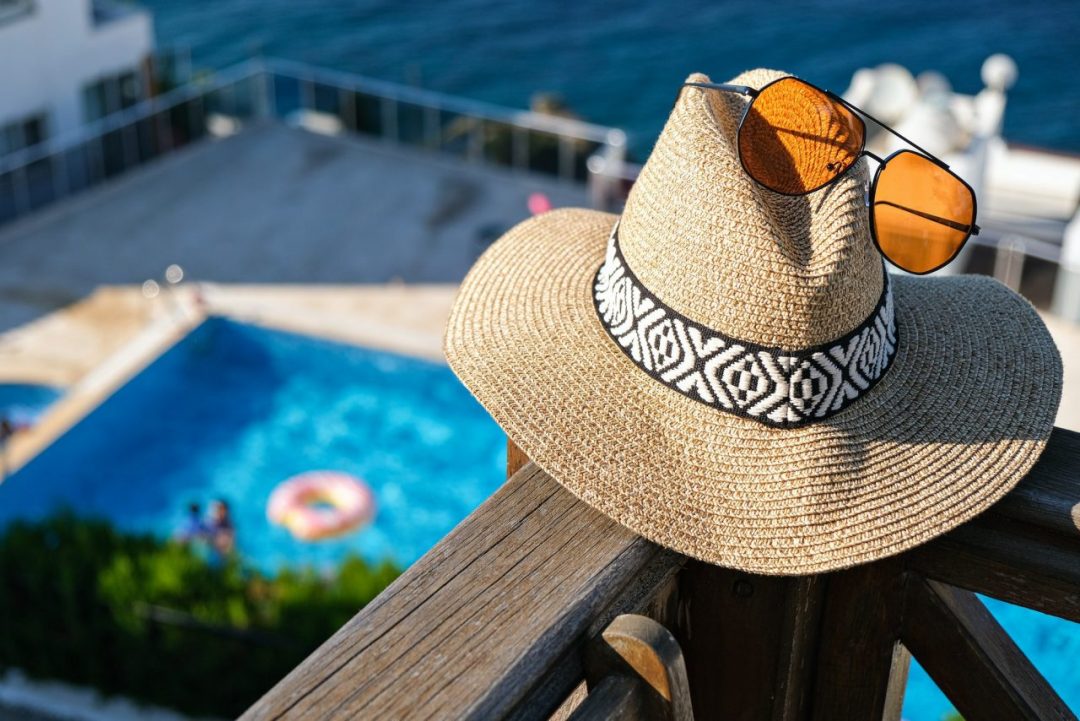 straw-hat-with-sunglasses-wooden-terrace-of-holiday-villa-or-hotel-with-sea-and-wimming-pool-view-e1673186375233